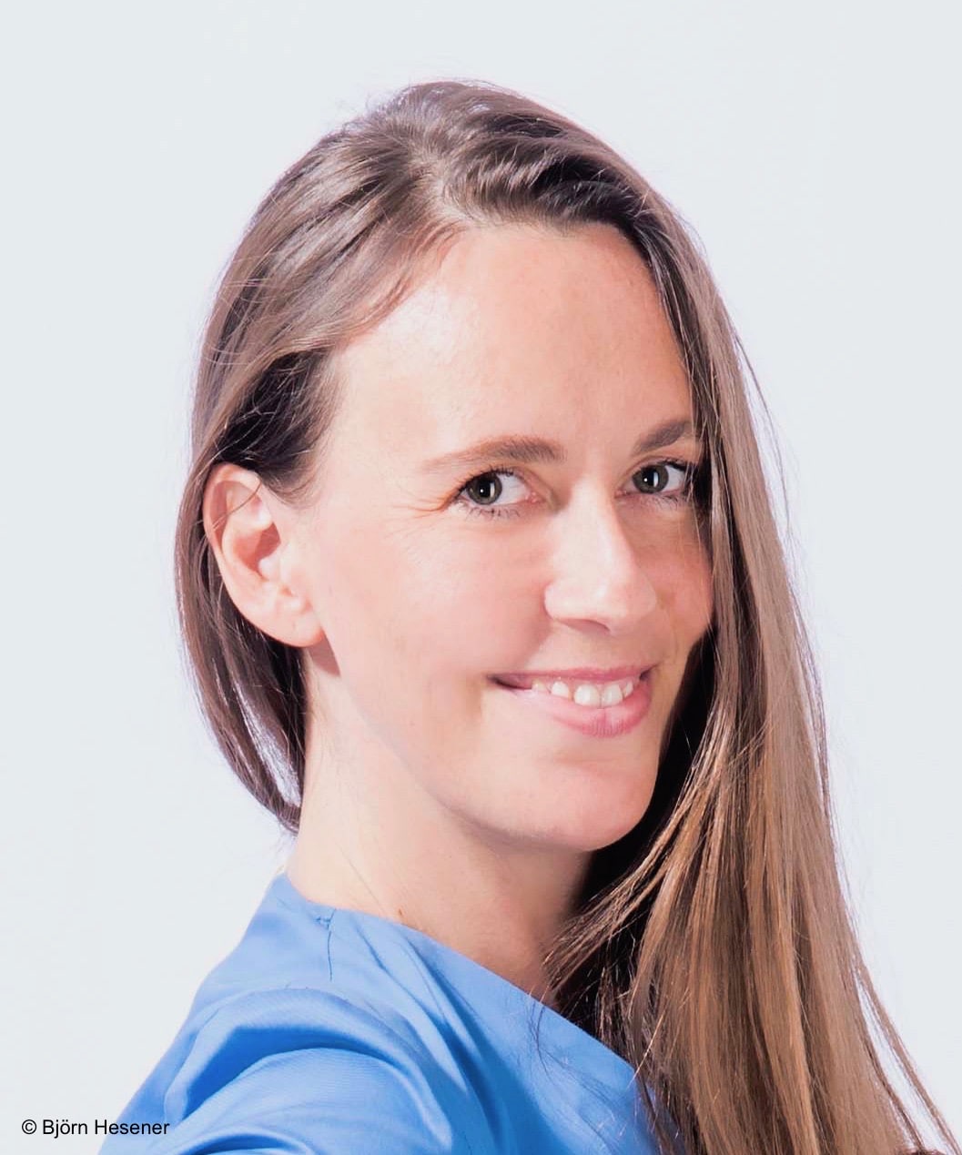 Frau mit langen braunen Haaren lächelt, trägt hellblaues Shirt, vor hellem, neutralem Hintergrund.