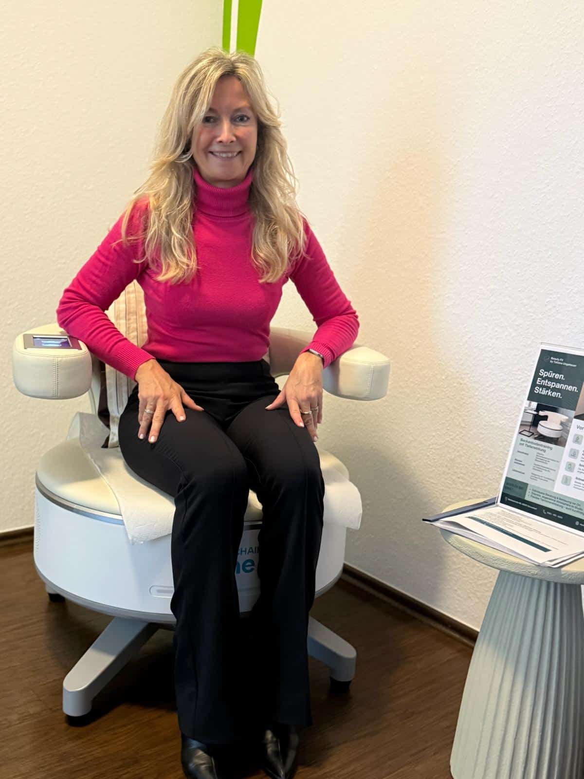 Frau mit langen blonden Haaren im rosa Rollkragenpullover sitzt lächelnd auf weißem Behandlungsstuhl.