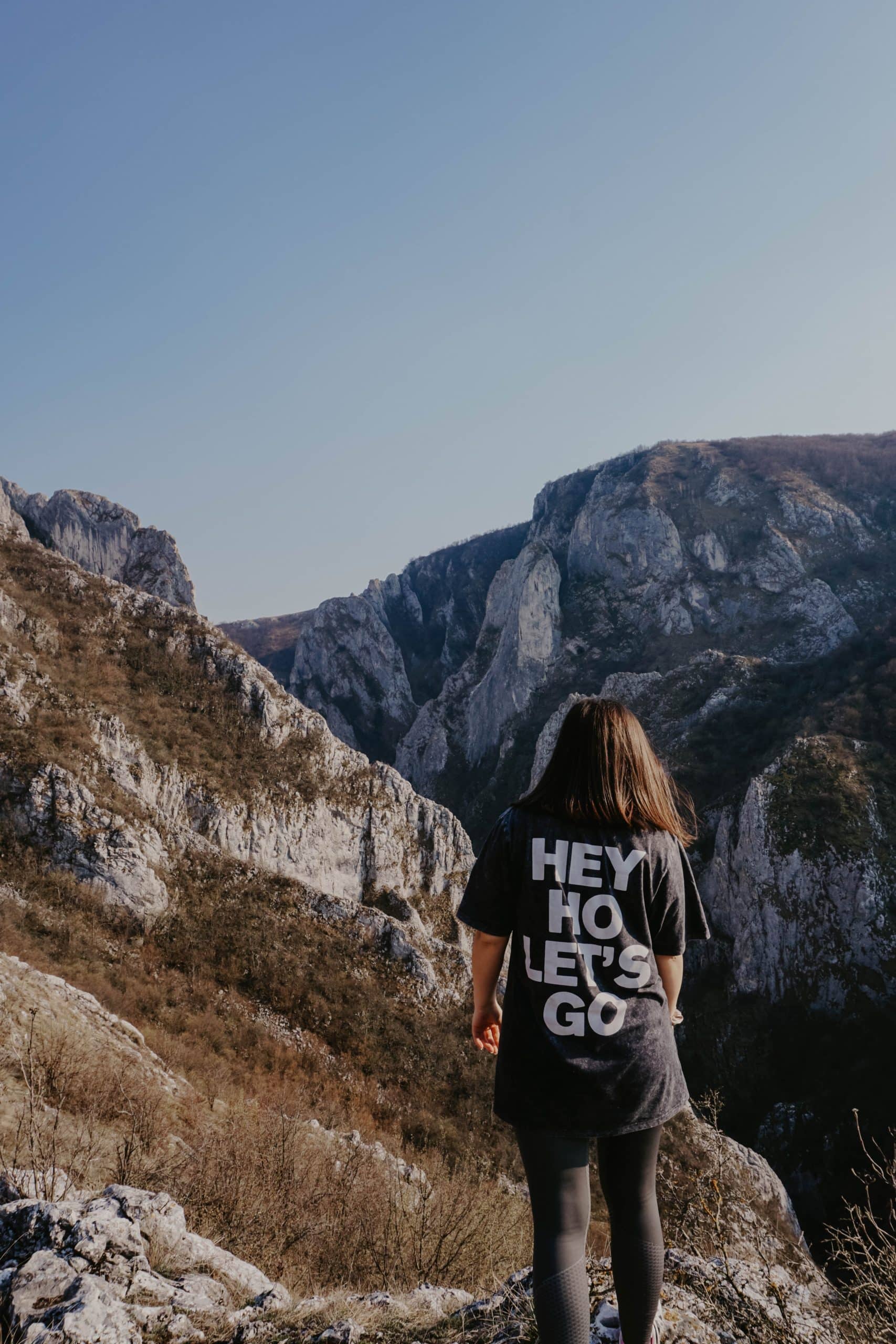 Frau im "HEY HO LET'S GO"-Shirt steht auf felsigem Gelände und blickt zu entfernten Bergen unter blauem Himmel.