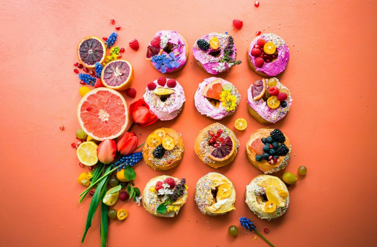 Bunte Donuts mit Früchten und Blumen liegen nebeneinander auf orangefarbenem Untergrund.