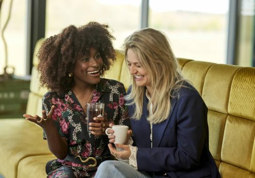 Zwei Frauen sitzen lachend auf einem gelben Sofa und trinken gemeinsam, während sie Essen und Lifestyle genießen.