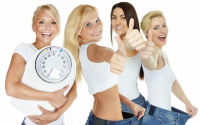 Vier Frauen in Jeans und weißen Oberteilen feiern mit Vitalstoffriegeln, zwei halten weite Jeans, eine eine Waage.