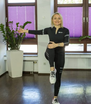 Frau in Sportkleidung macht im Büro eine aktive Bewegungspause und blickt dabei lächelnd in die Kamera.