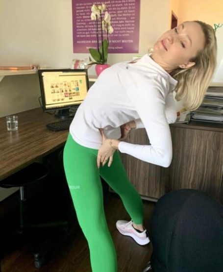 Frau in grüner Leggings macht Dehnübungen am Schreibtisch mit Computer und Orchidee im Büro.