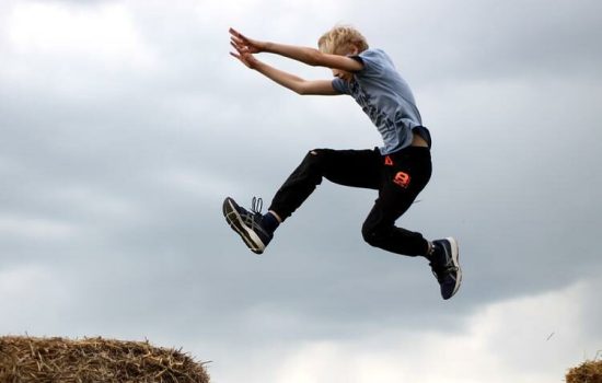Junge springt in einem Präventionskurs für Grundschulkinder zwischen zwei Strohballen unter bewölktem Himmel.