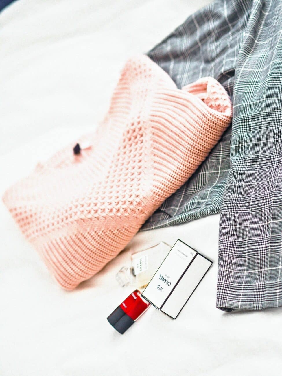 Rosa Strickpullover, karierte Hose und Parfümprobe ausgebreitet auf einer Fläche.