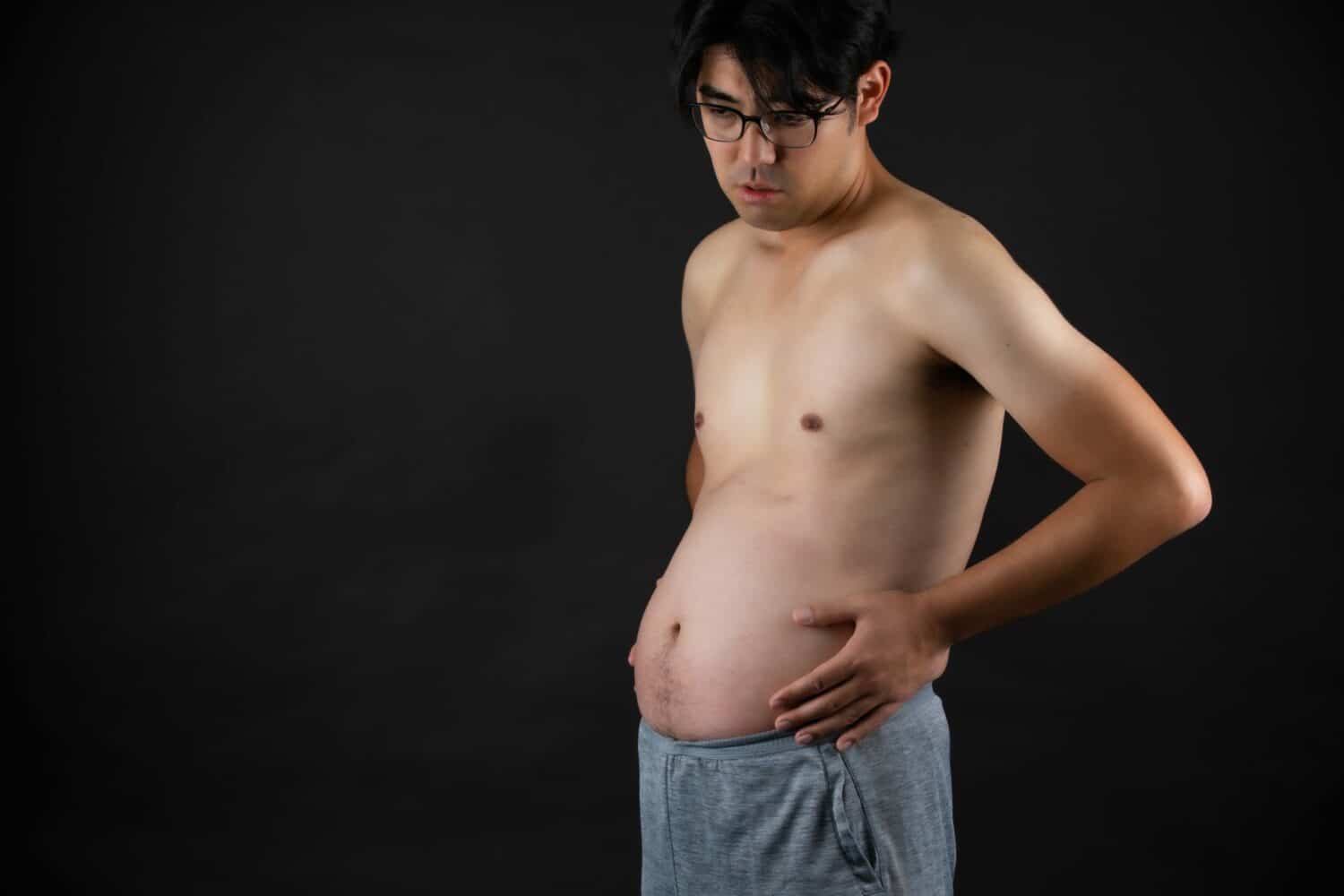 Mann ohne Hemd und mit Brille betrachtet seinen aufgeblähten Bauch, im Zusammenhang mit My Beautyfit-Kleidung.